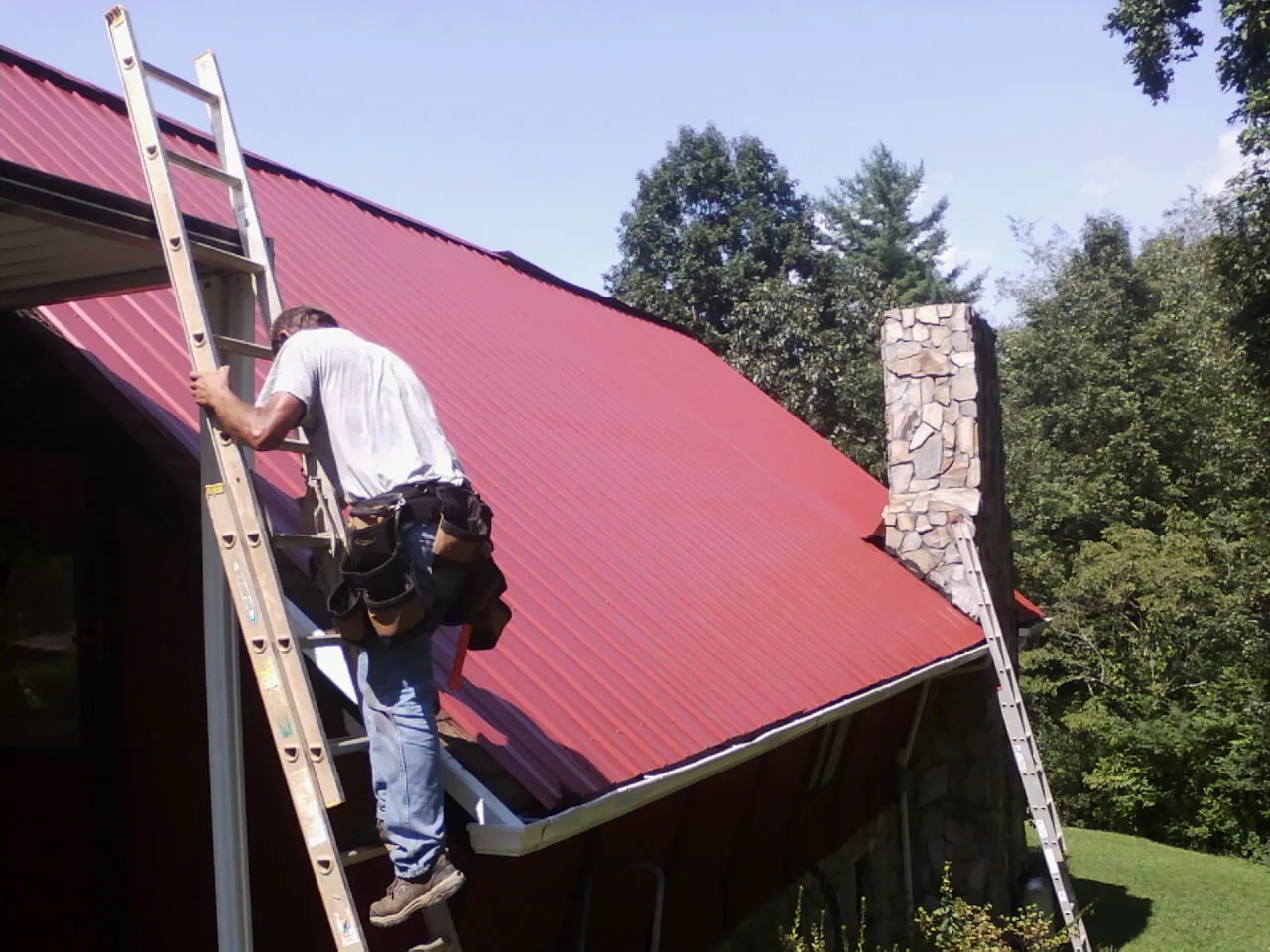 Roofer on a ladder beside a red metal roof and stone chimney — Soffit & Fascia Repair by Roofing Brothers in Haugan